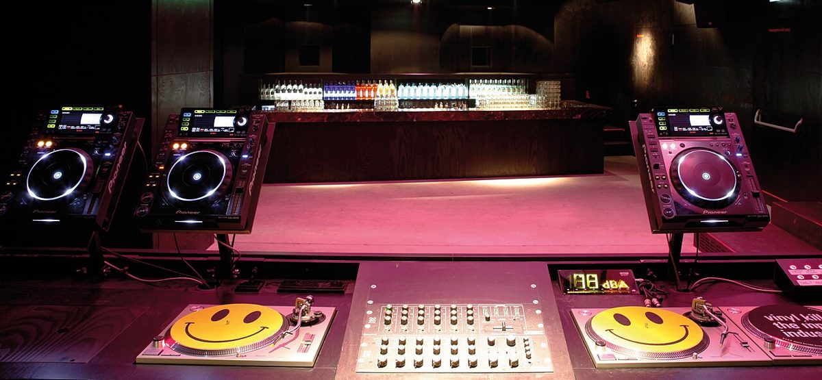 A DJ performing behind the booth with crowd and lights in a nightclub