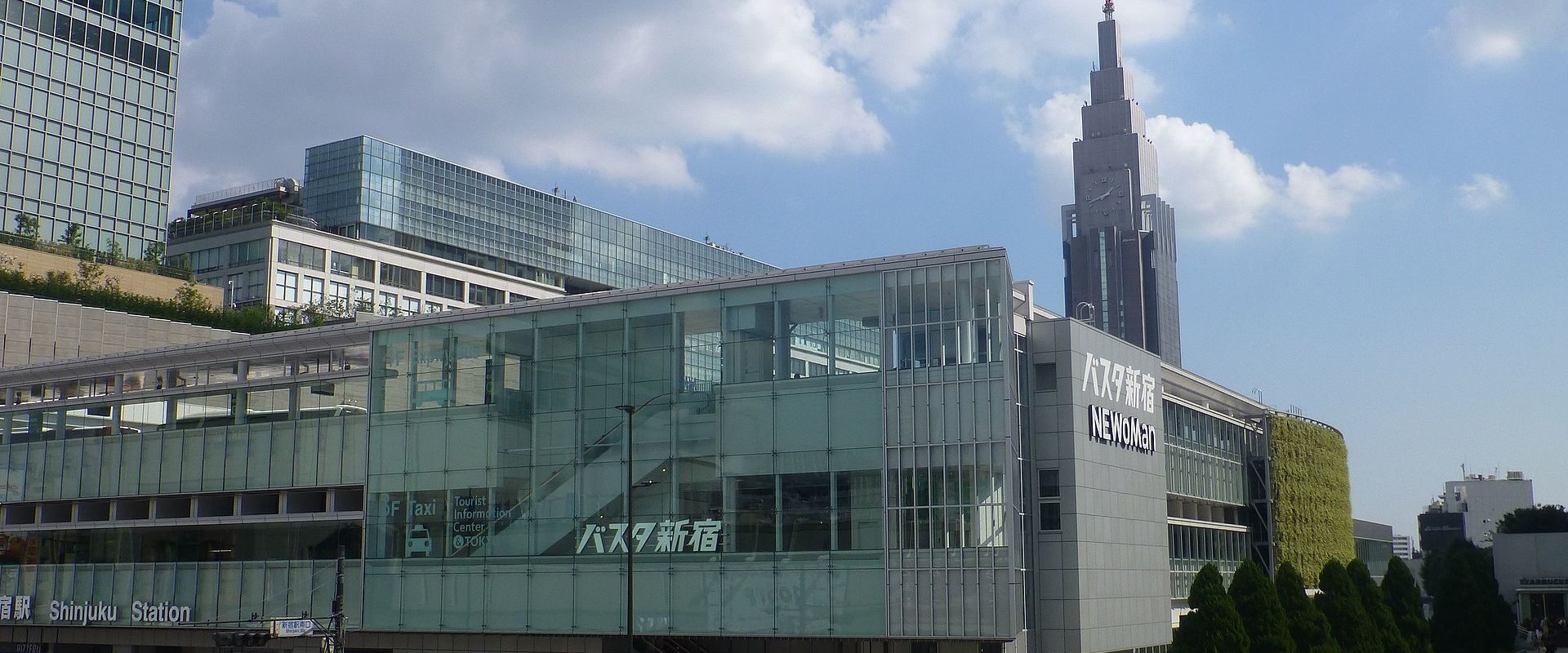 The glass Busta Shinjuku terminal building above Shinjuku Station's New South Exit with crowds on the street and the Docomo tower behind