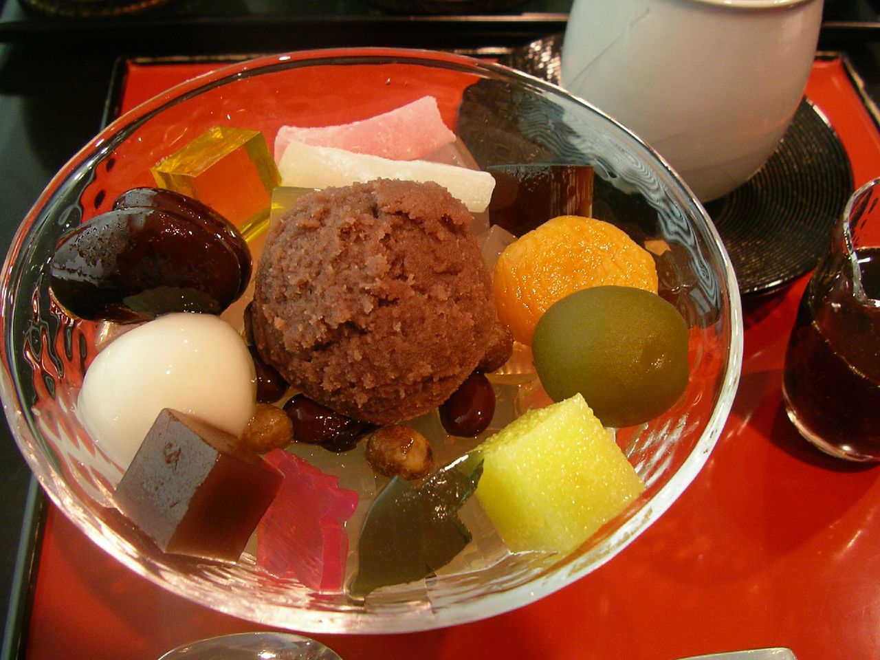 A glass bowl of cream anmitsu with red bean paste, agar jelly, mochi, fruit and a scoop of ice cream on a red lacquer tray