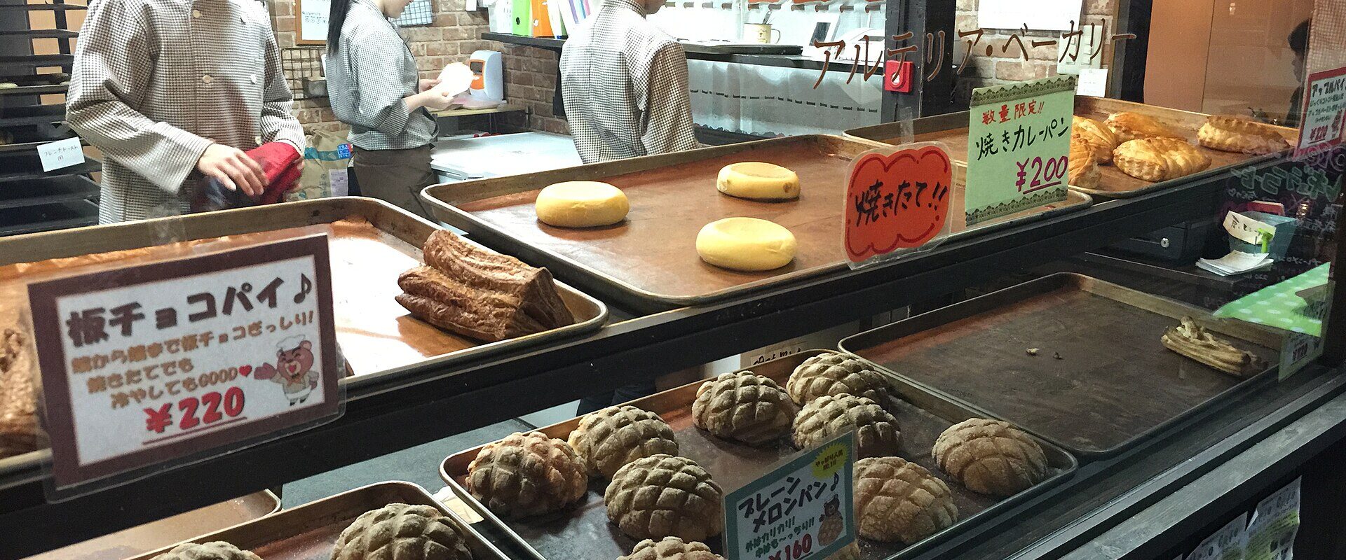 Bakers in white caps working behind a counter of crosshatched melon pan and chocolate buns at a Japanese bakery