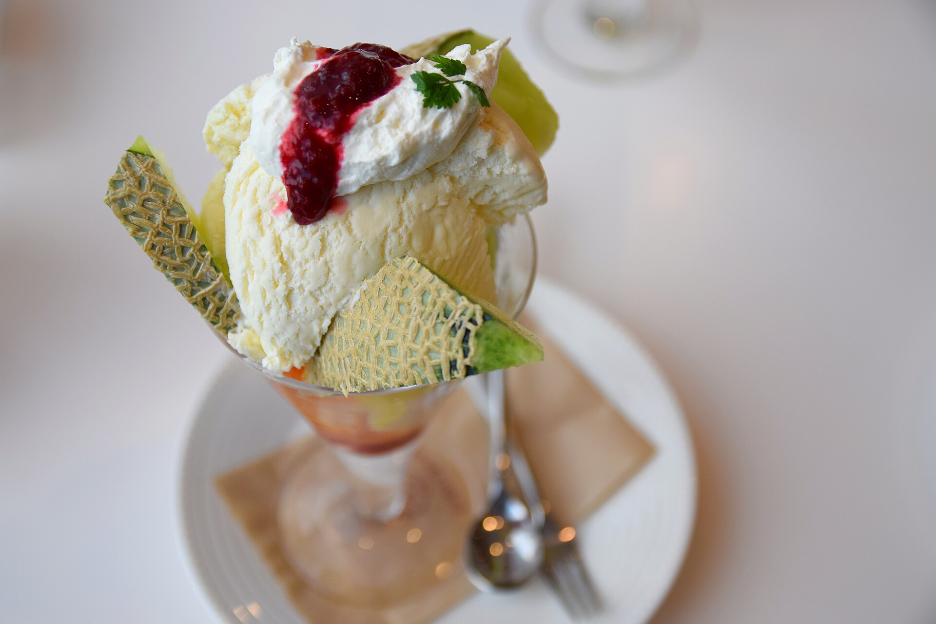 A tall fruit parfait topped with wedges of green melon, whipped cream and berry sauce