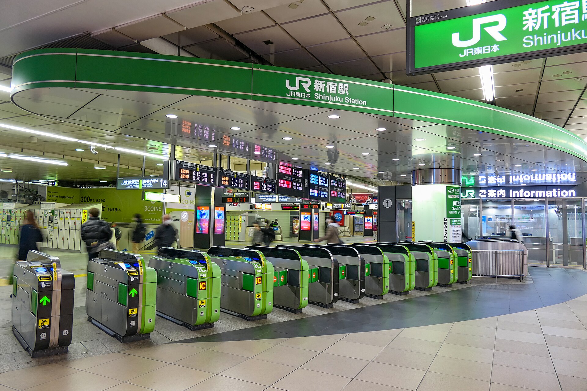 A row of green JR ticket gates beneath the curved Shinjuku Station sign at the South Gate, with departure boards behind