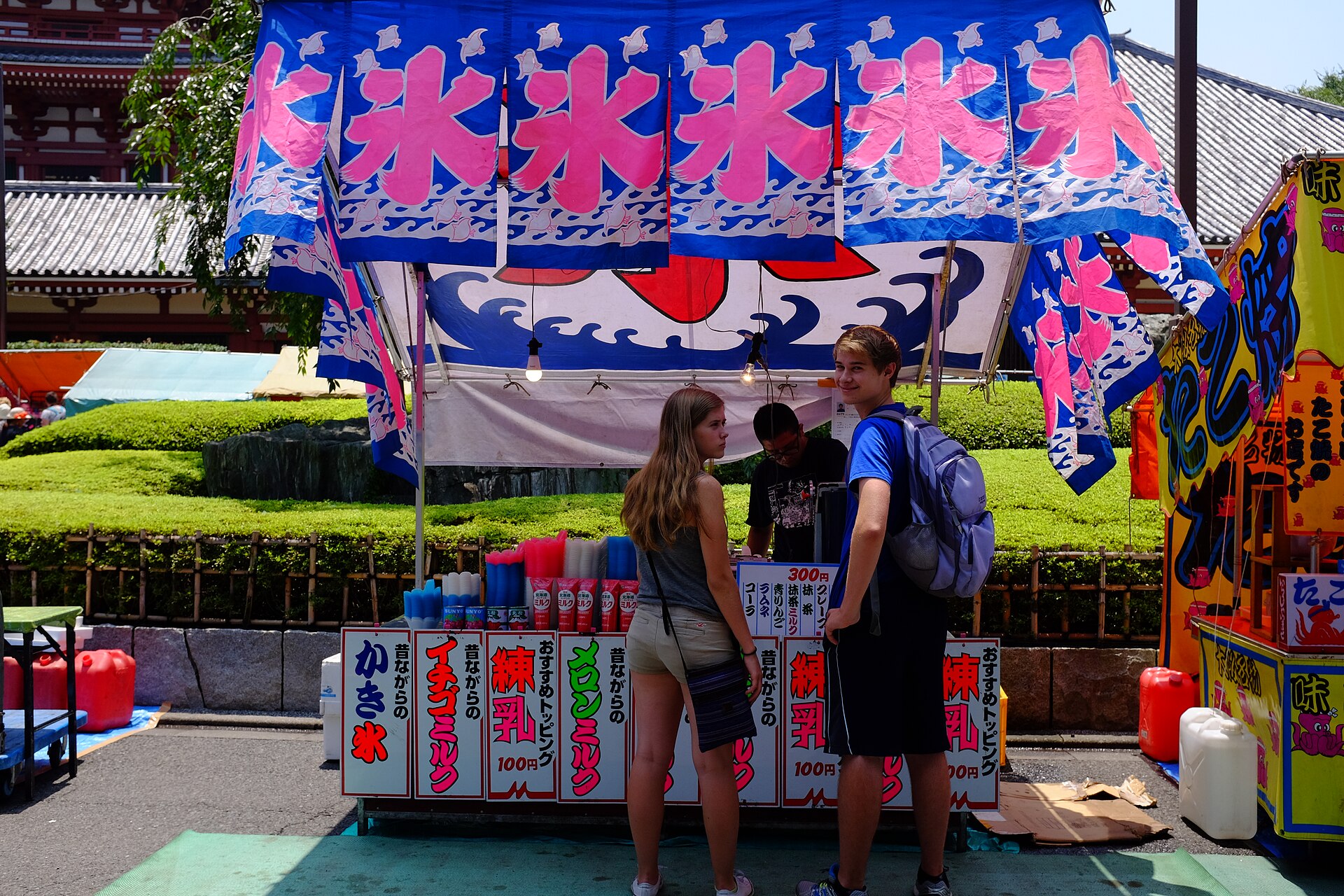 Visitors buying shaved ice at a festival stall with blue and red ice flags in front of a temple