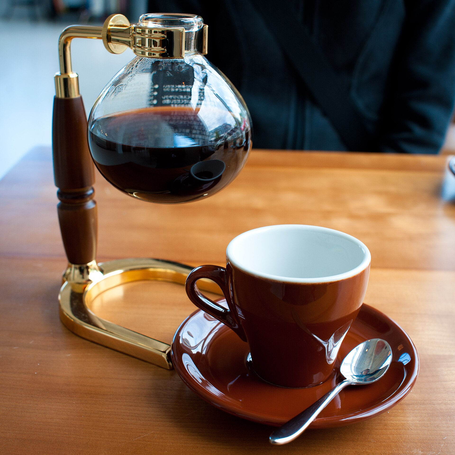 A glass siphon coffee brewer on a wooden table beside a cup of black coffee