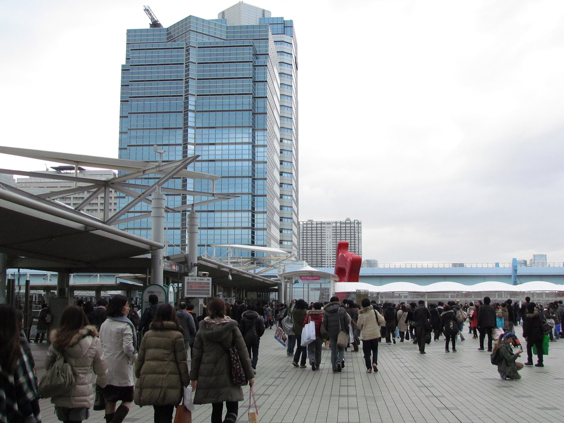 Crowds of Comic Market visitors in winter coats carrying shopping bags across the plaza outside Tokyo Big Sight, with an office tower behind