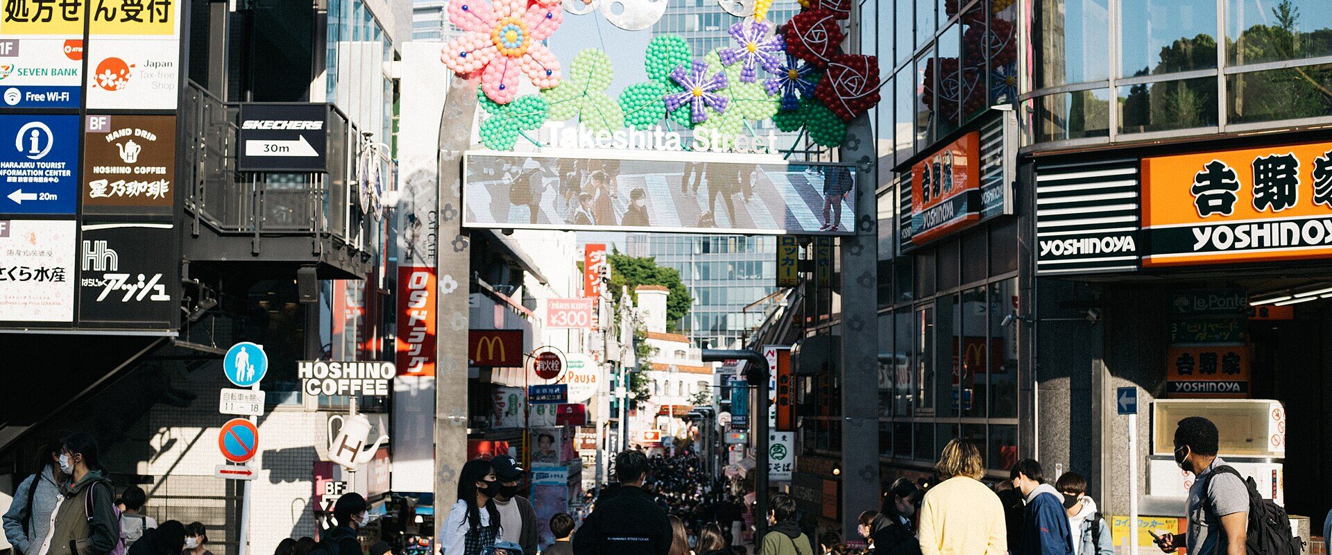 Crowds enter Takeshita Street in Harajuku, Tokyo, beneath an arch decorated with colorful flowers and a street sign.