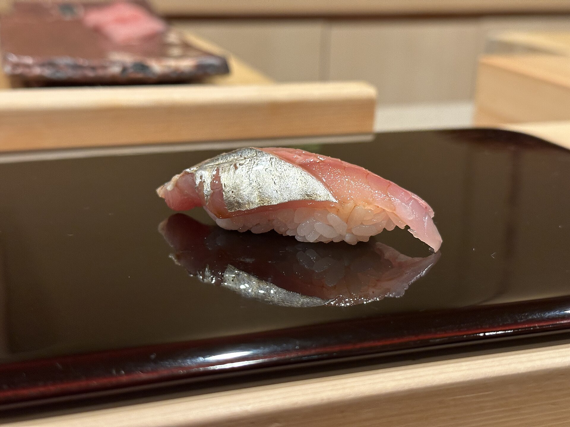 Horse-mackerel nigiri with silvery skin rests on a glossy dark plate at a wooden sushi counter in Tokyo.