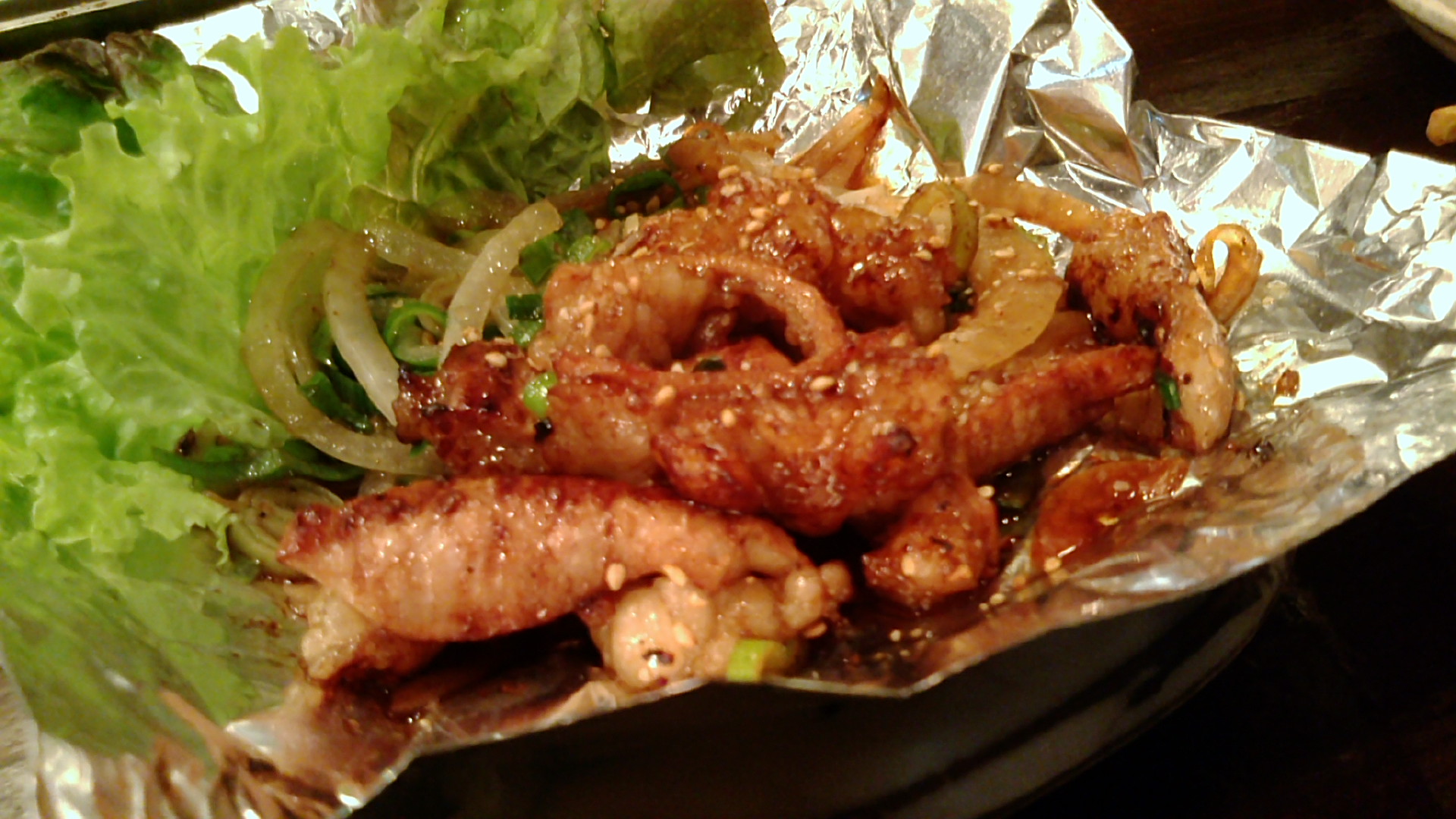 Grilled horumon offal glazed with sauce and sesame seeds with onions on foil, beside lettuce leaves