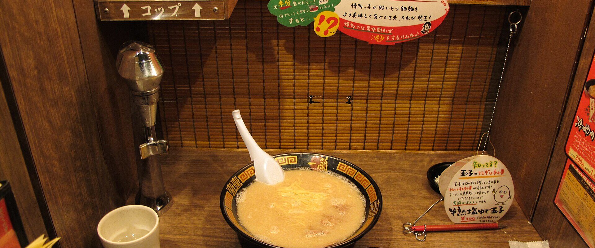 A bowl of tonkotsu ramen and a boiled egg in a wooden single-seat booth at Ichiran, with a water tap and order sheet