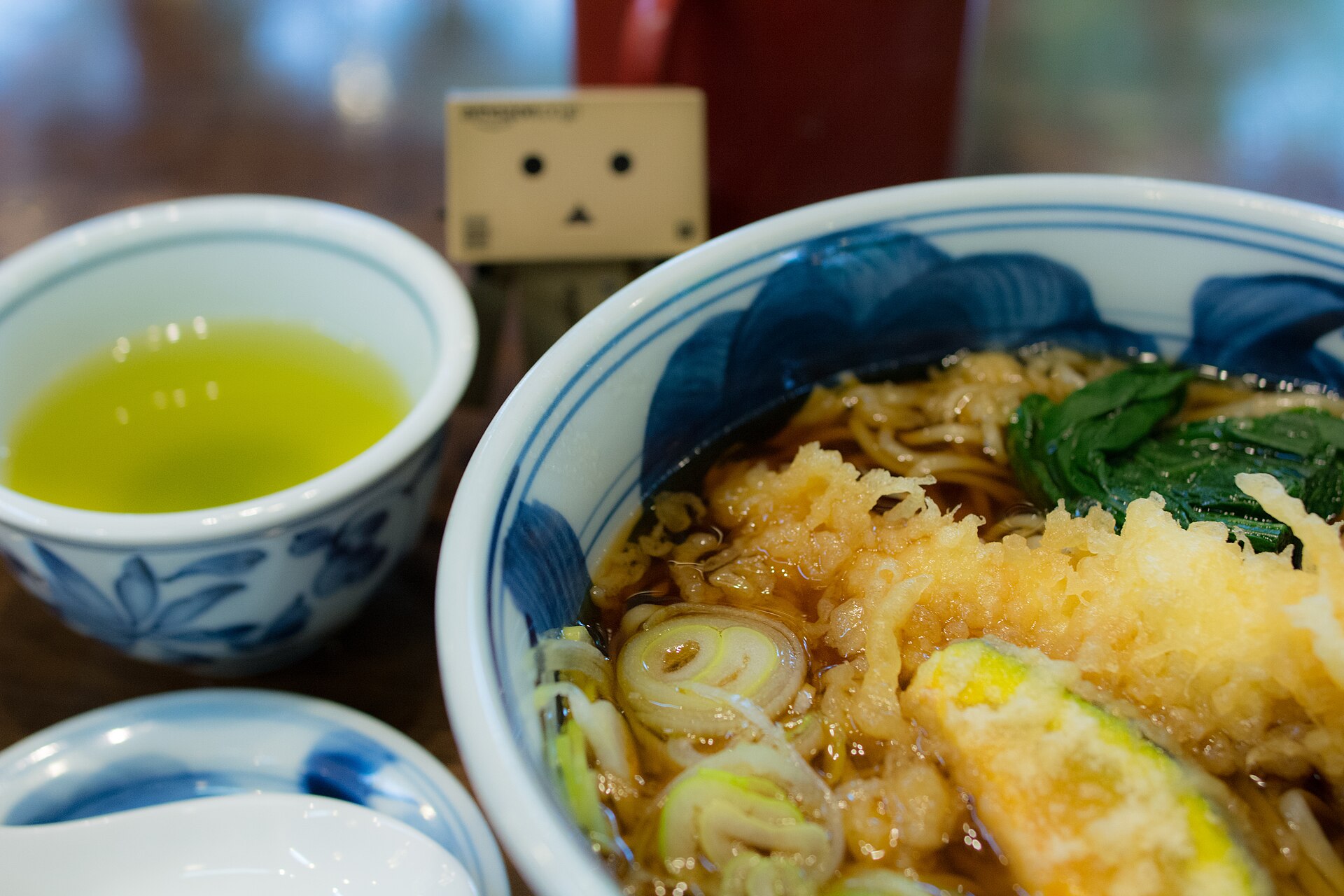 A bowl of hot tempura soba with spring onion and greens beside a cup of green tea at a restaurant near Jindaiji temple