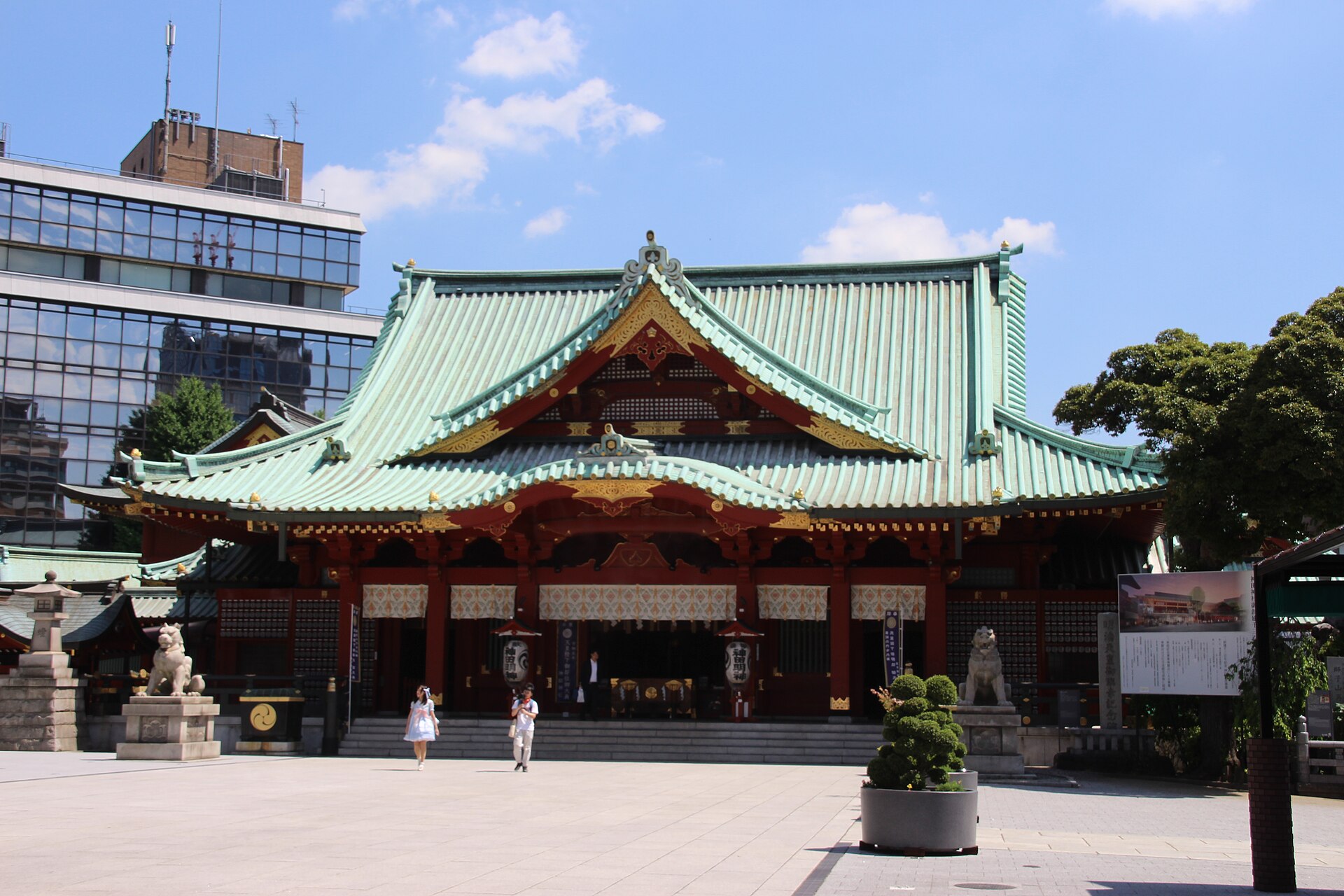 The red and green main hall of Kanda Myojin shrine under a blue sky, with a paved courtyard and stone guardian lions