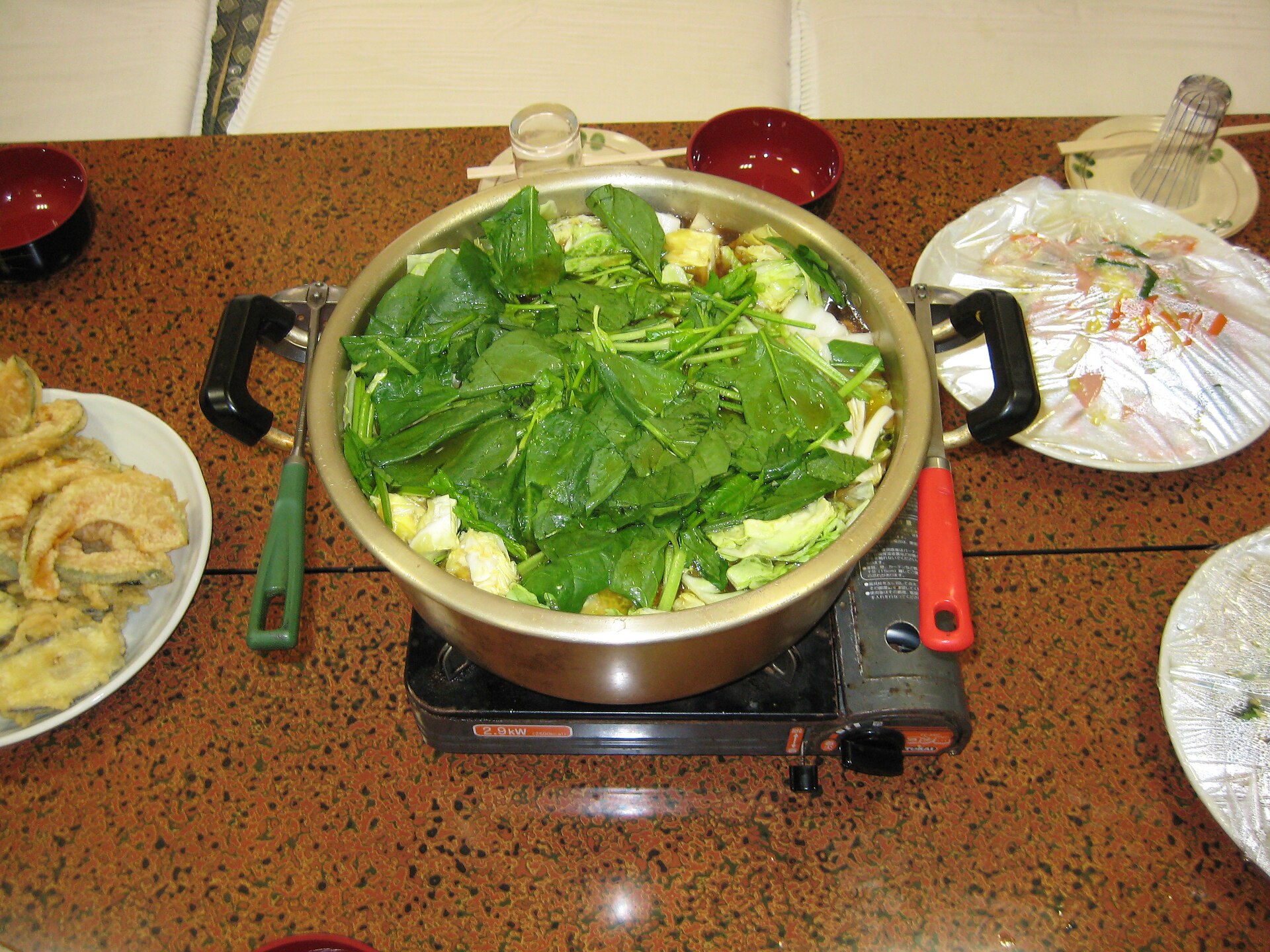 A large chanko nabe pot filled with leafy greens sits on a tabletop burner, surrounded by bowls and side dishes.