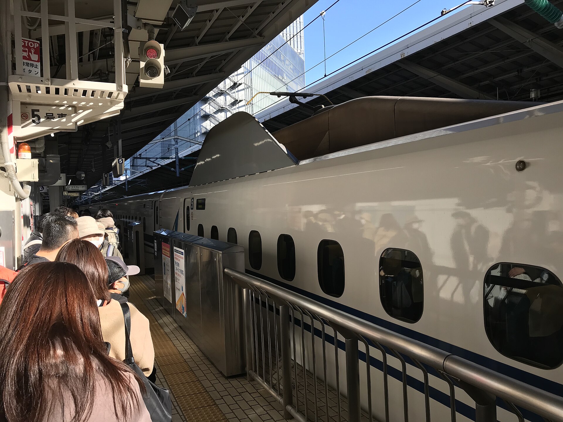 Passengers queue beside a white and blue Shinkansen at Tokyo Station, with low platform barriers separating them from the train.