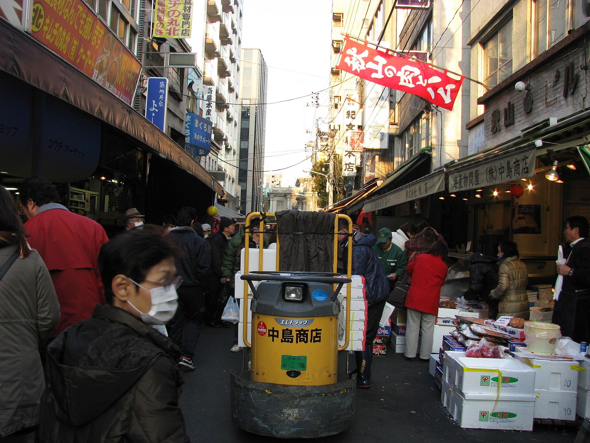 Shoppers and a small delivery vehicle share a narrow lane beside open seafood boxes at Tsukiji Outer Market in Tokyo.