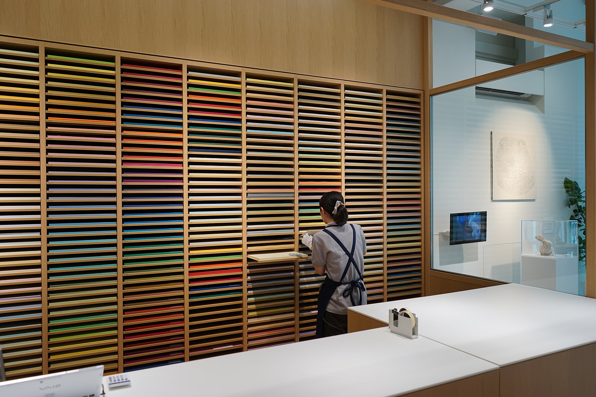A staff member stands beside colourful paper sheets arranged in shallow wooden shelves at Ito-ya in Ginza, Tokyo.