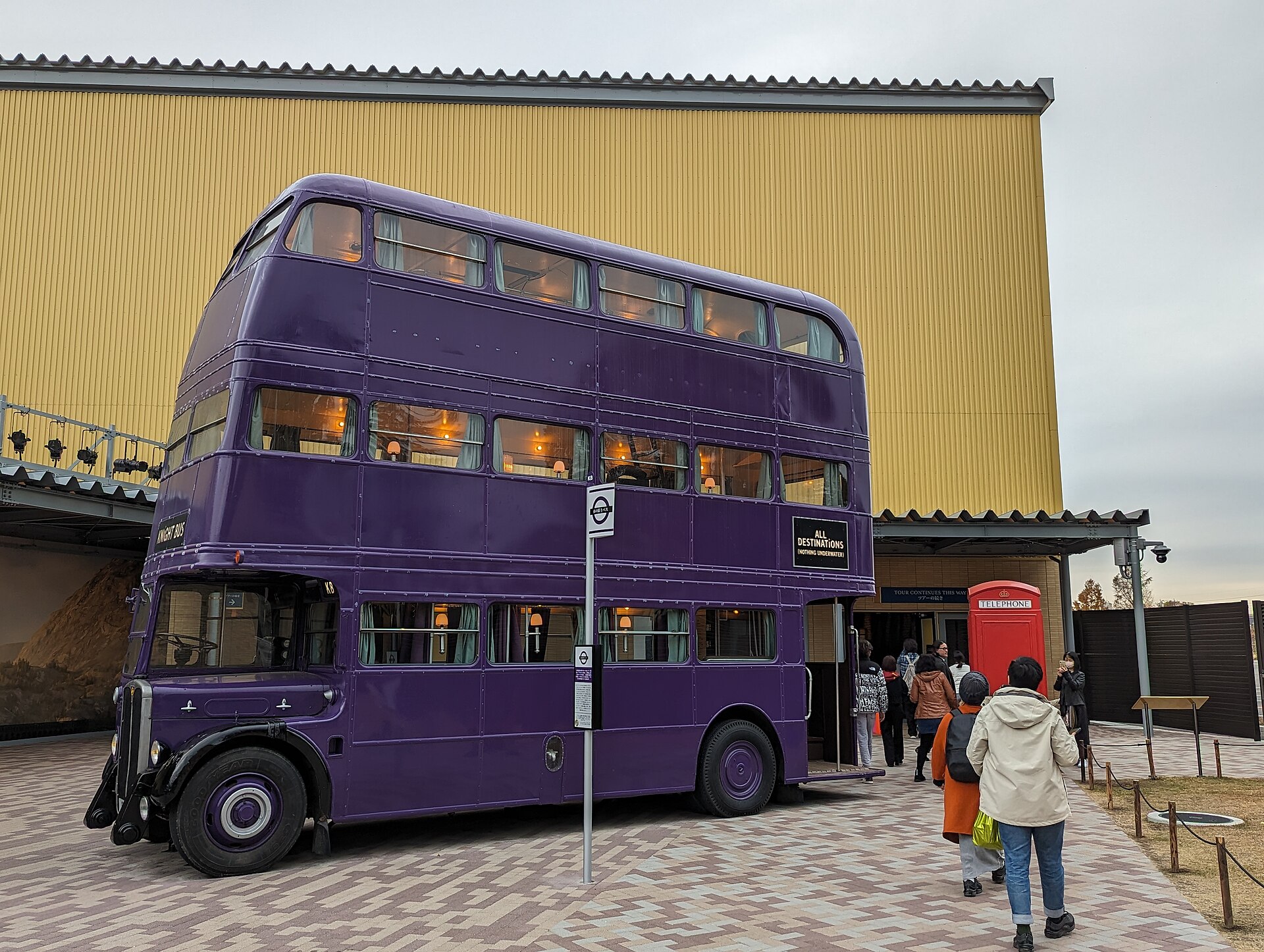 The purple triple-decker Knight Bus parked outside a yellow studio building, with visitors walking past a red telephone box