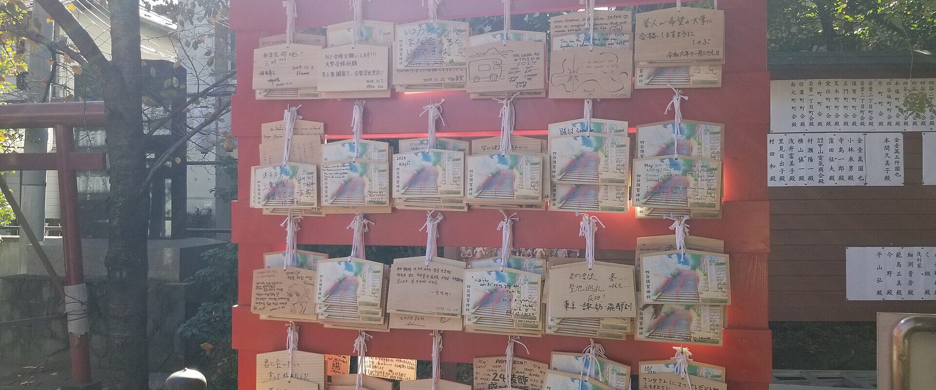 Rows of wooden ema prayer plaques illustrated with the Suga Shrine staircase, covered in handwritten wishes from visitors, on a red rack at Suga Shrine