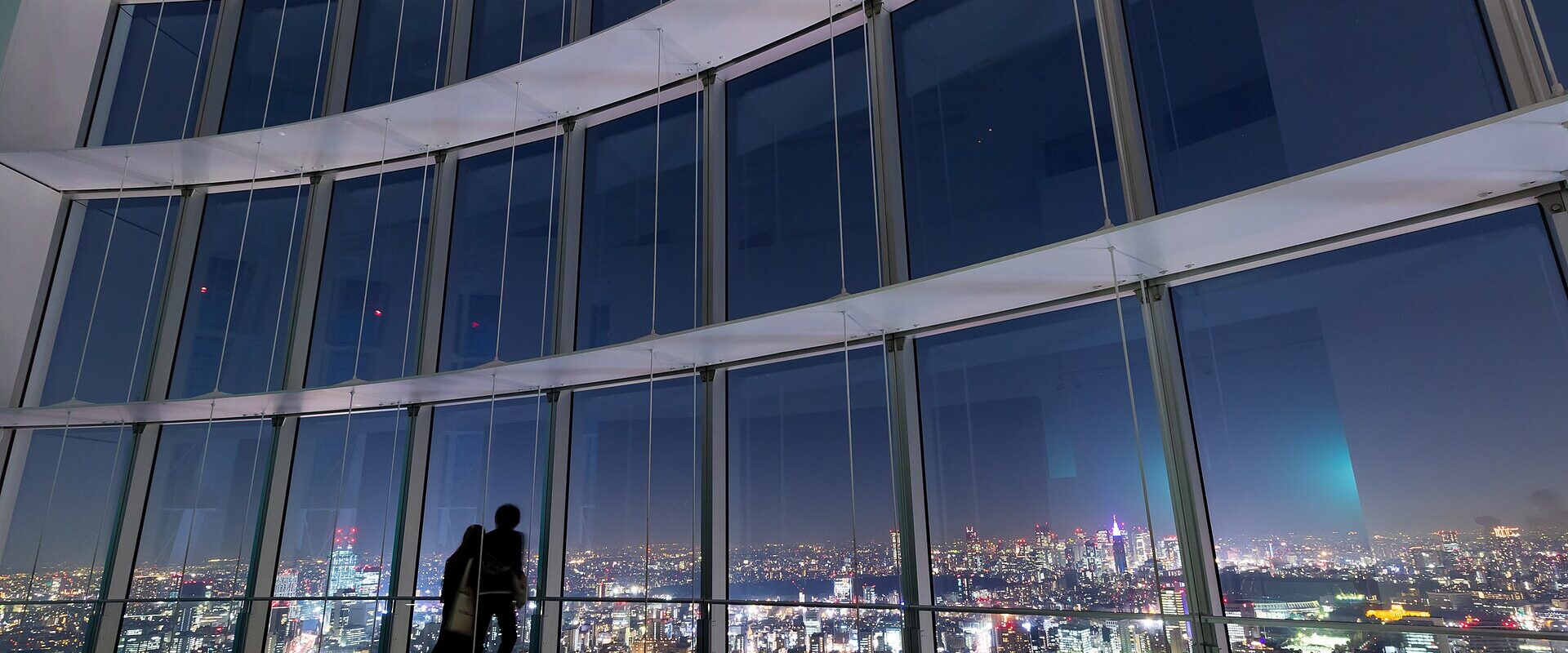 Two visitors silhouetted against the tall windows of Tokyo City View at night, with the lit-up city spread out below