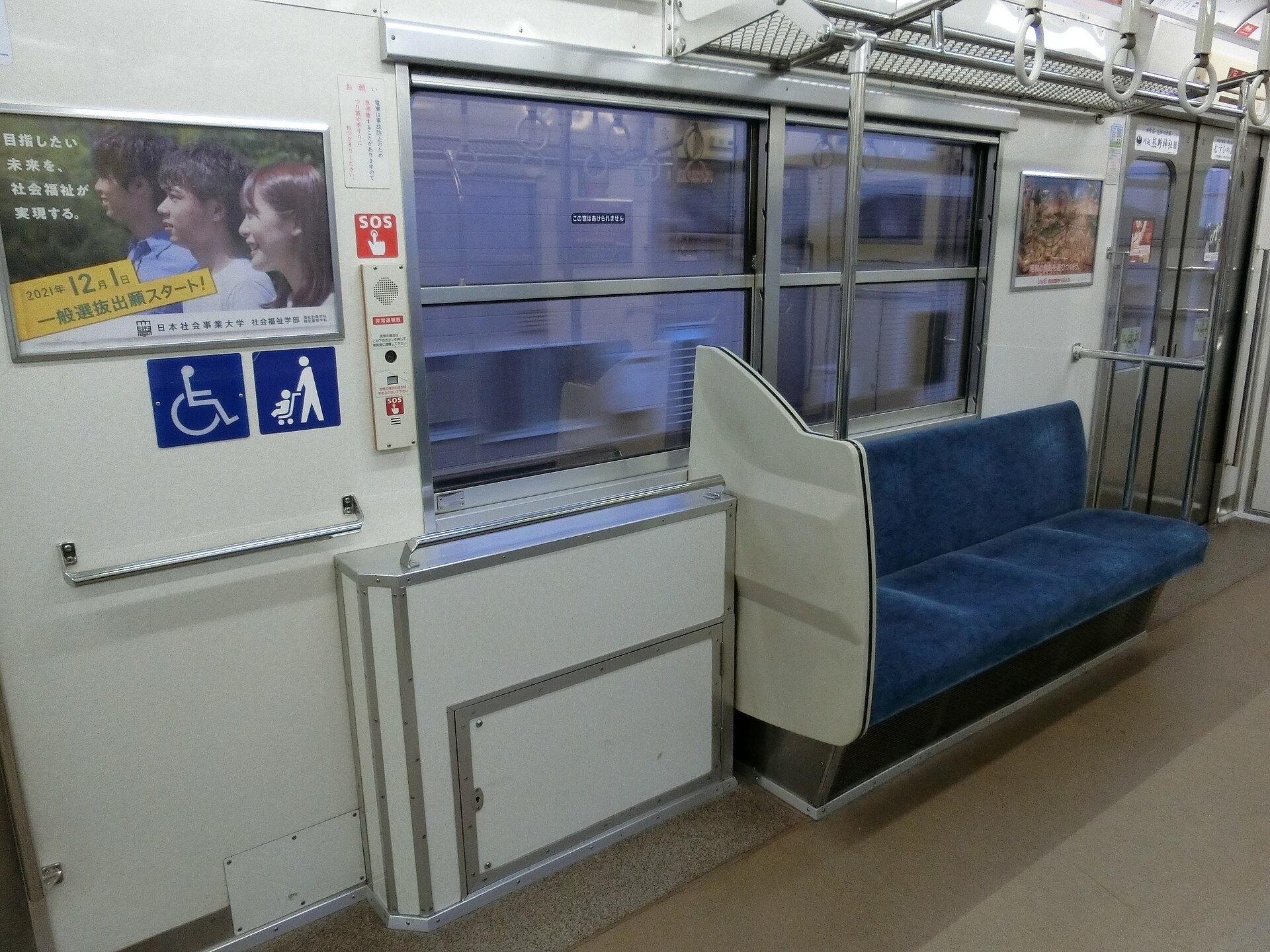 A wheelchair space inside a Japanese commuter train, with a handrail, emergency call button and wheelchair and pushchair symbols on the wall