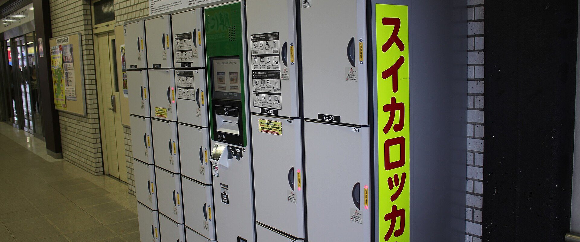 A bank of white coin lockers in a Japanese station with a central touch-screen payment panel and a yellow sign reading Suica locker