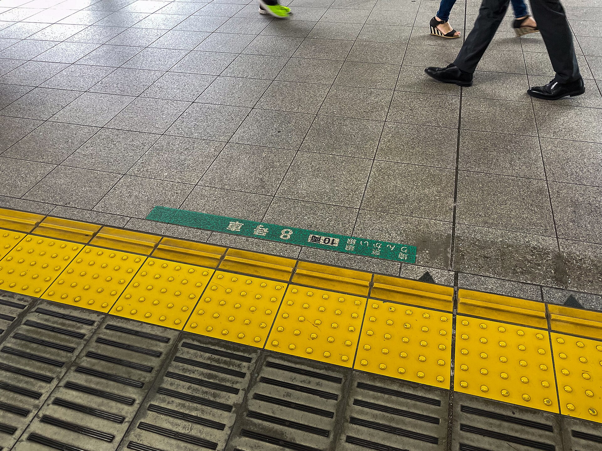 Yellow tactile paving with raised dots running along the edge of a Tokyo train platform, with commuters walking behind