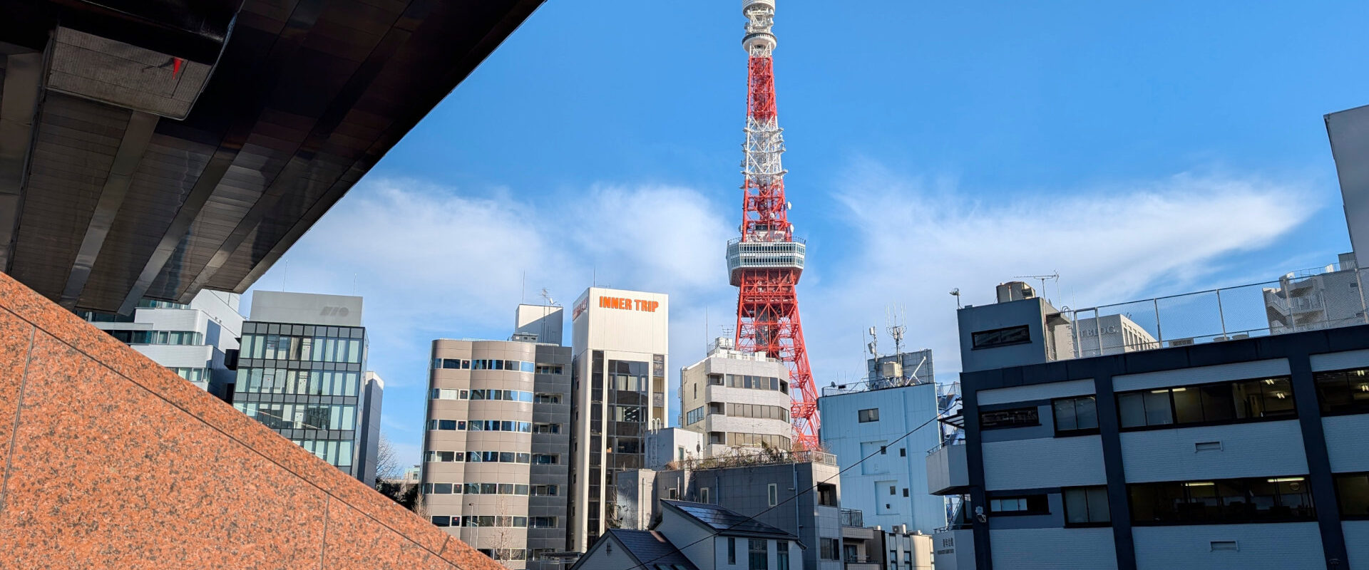 Tokyo Tower rises above nearby city buildings beneath a blue sky, framed by a dark overhang and stone wall.