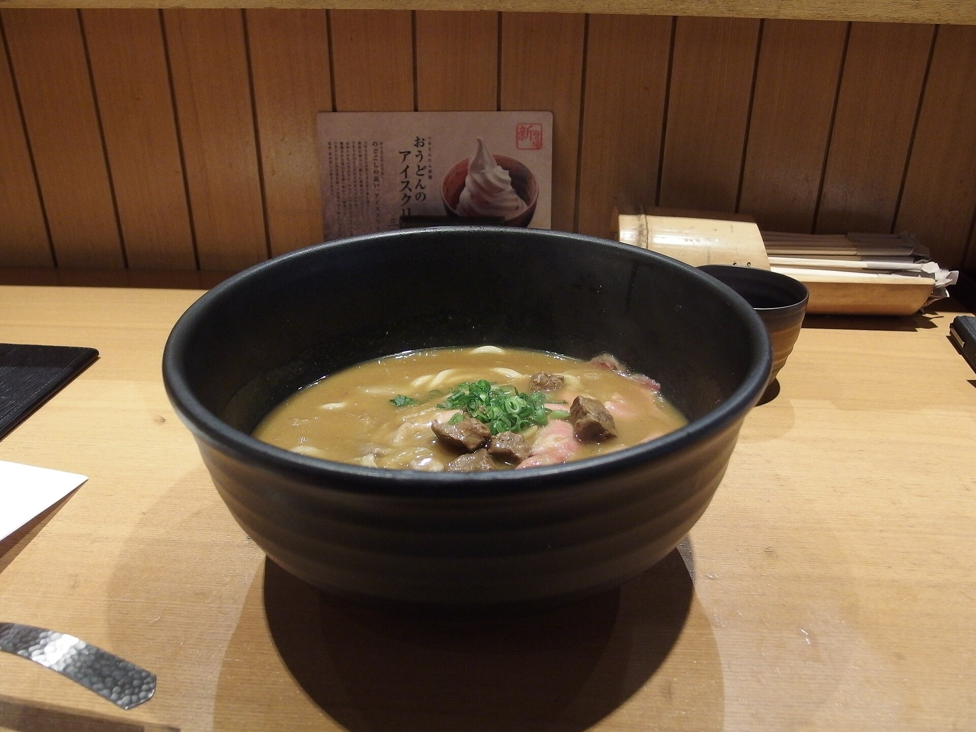 A large black bowl of curry udon with beef pieces and chopped spring onion on a wooden counter at Tsurutontan in Roppongi