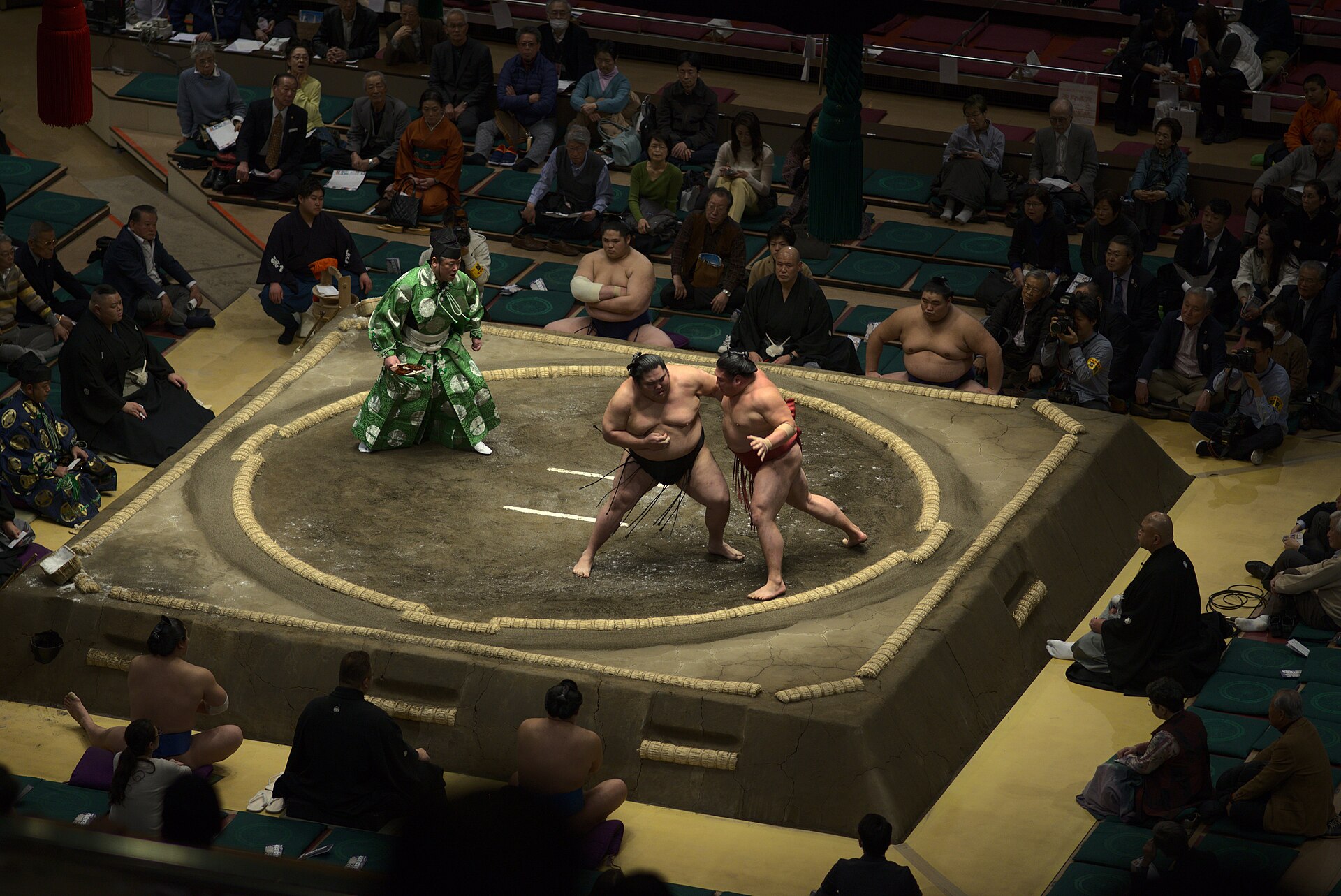 Two rikishi grapple on the dohyo at Ryogoku Kokugikan in Tokyo, watched by a referee and seated spectators.