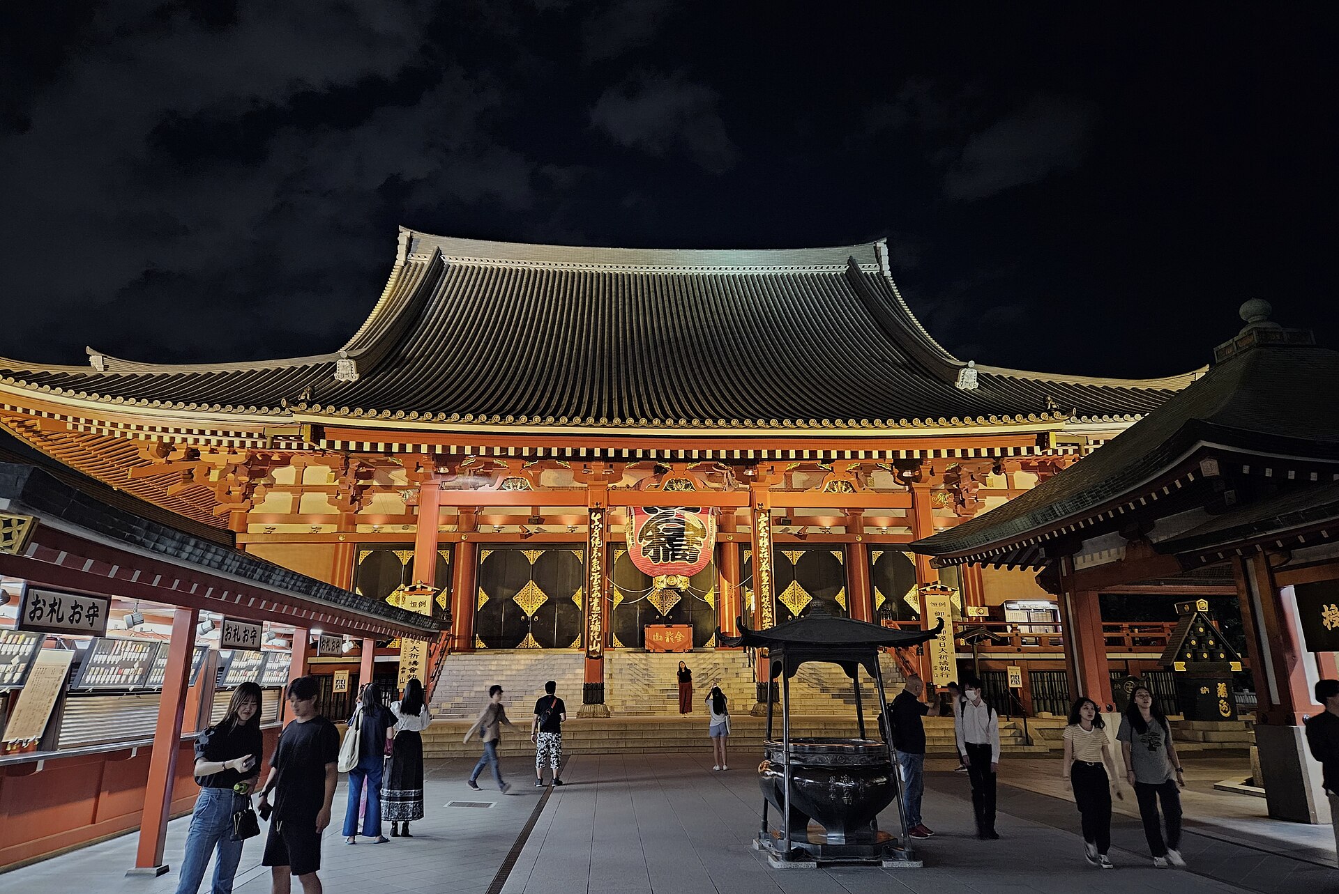 Visitors gather around the incense burner beneath the illuminated main hall of Senso-ji in Asakusa, Tokyo, after dark.