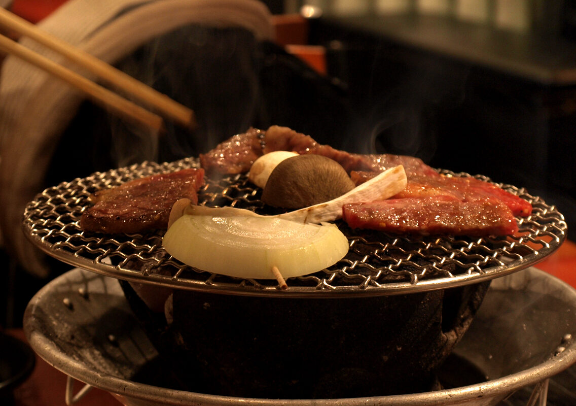 Slices of marbled beef, onion and mushrooms grilling over charcoal on a small round tabletop grill