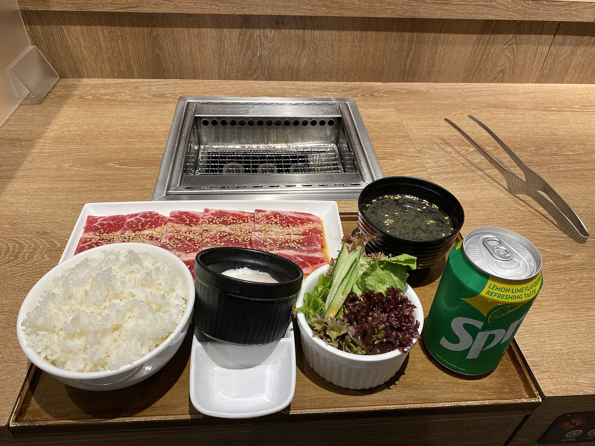 A Yakiniku Like one-person set of marbled beef slices, rice, soup, salad and an egg in front of a small individual grill at a counter